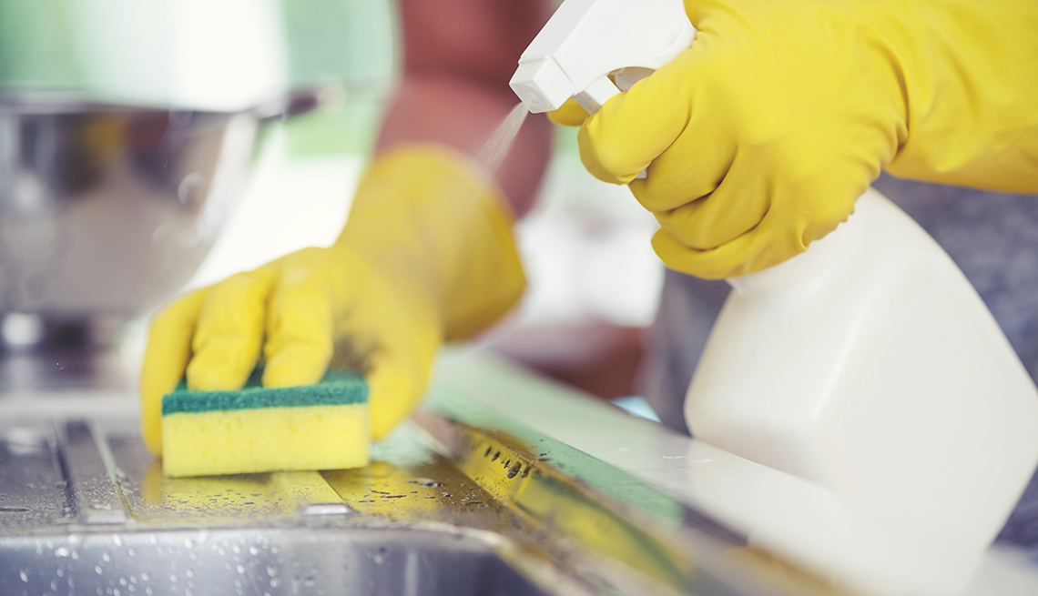 person wearing rubber gloves using a spray bottle and sponger to clean a kitchen sink