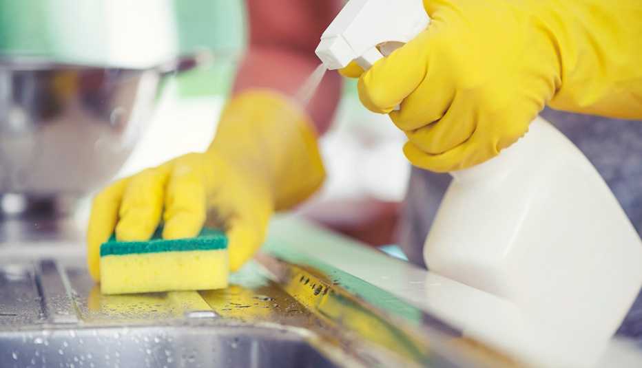 person wearing rubber gloves using a spray bottle and sponger to clean a kitchen sink