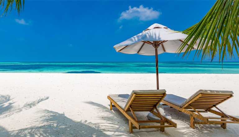 two chairs on white sand beach, turquoise water, with umbrella and palms from a tree on side