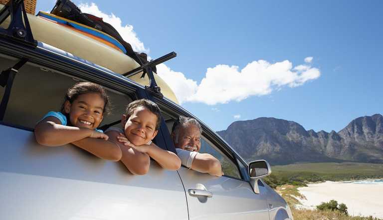passenger side of a car with luggage on the roof rack, mountains in the background, blue skies and white clouds, grandfather looking out front seat passenger seat, two children, boy and girl, sitting with arms out of the back seat window