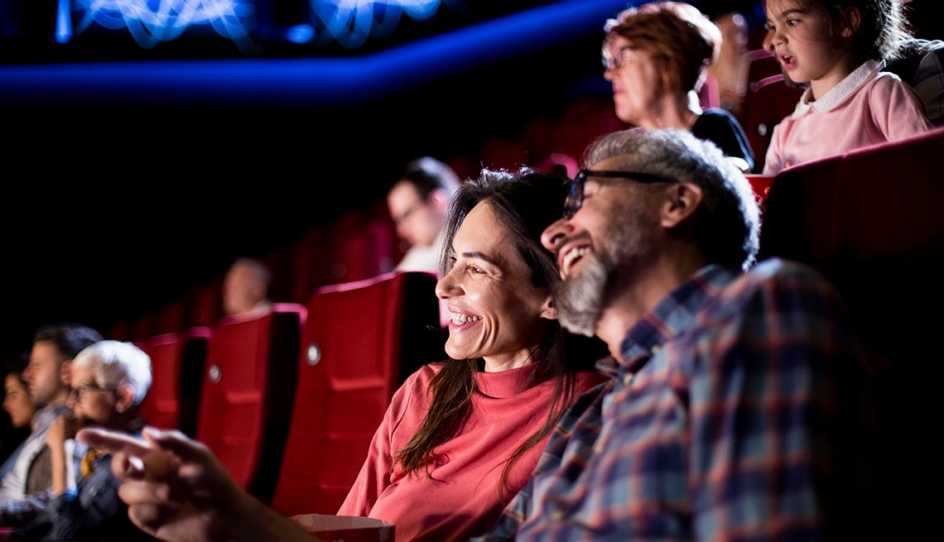 couple, woman and man sitting in movie theater, smiling, watching movie, other people around them in theater including small female child and grandma behind the couple