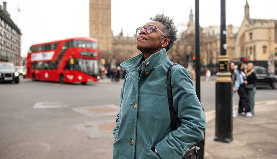 woman wearing glasses with green coat, looking up at sky in european city, red doubledecker bus in background