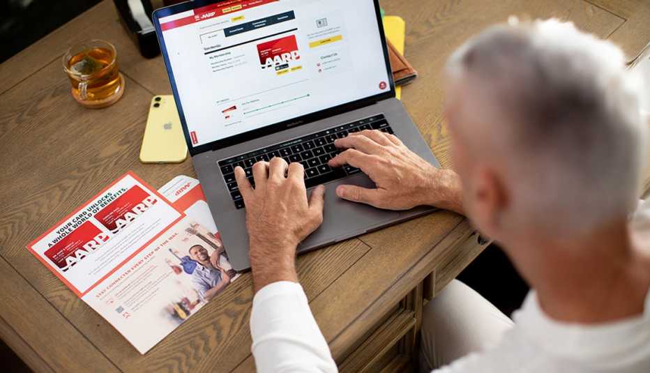 A man typing at his laptop sitting at a table visiting My Account on the AARP.org site while viewing his AARP new member Welcome Kit received in the mail.