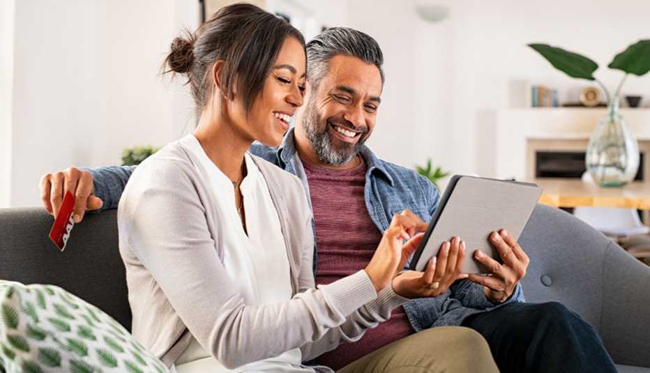 A man and woman sitting on a couch, looking at a tablet smiling.