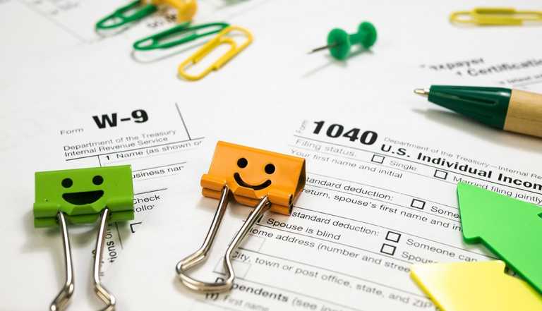 Orange and green binder clips with smiley faces sit on top of IRS tax forms including a 1040 and W-9. 