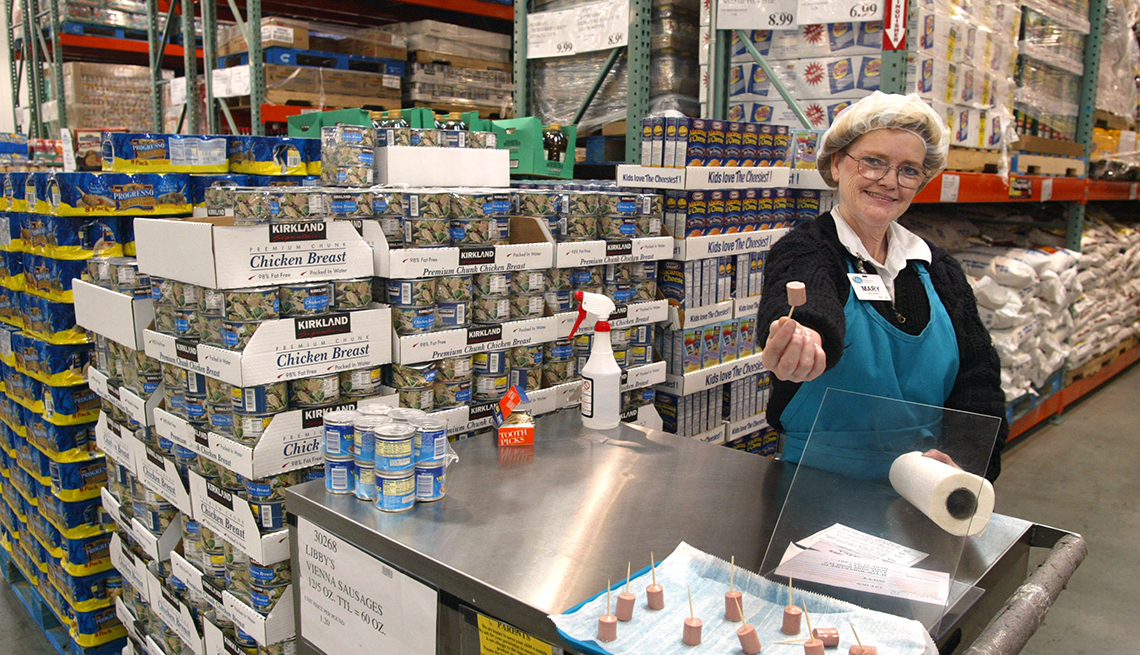 Costco warehouse worker with a tray of free samples on toothpicks, holding up sample for photo.