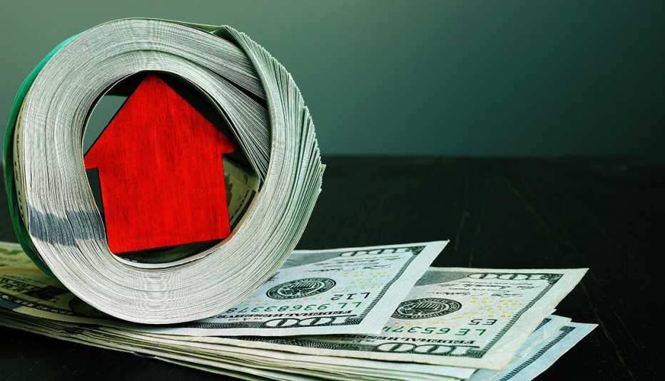 a minature wooden red house is wrapped inside a roll of hundred dollar bills on a dark green background
