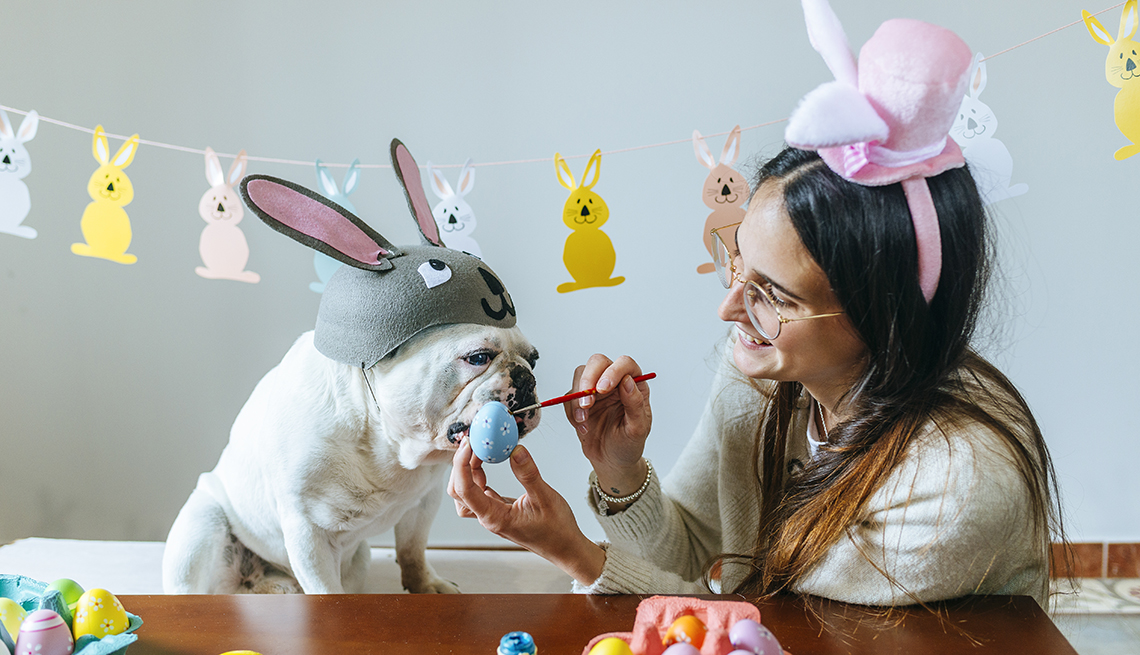 woman paints easter eggs while a bulldog in a bunny hat watches