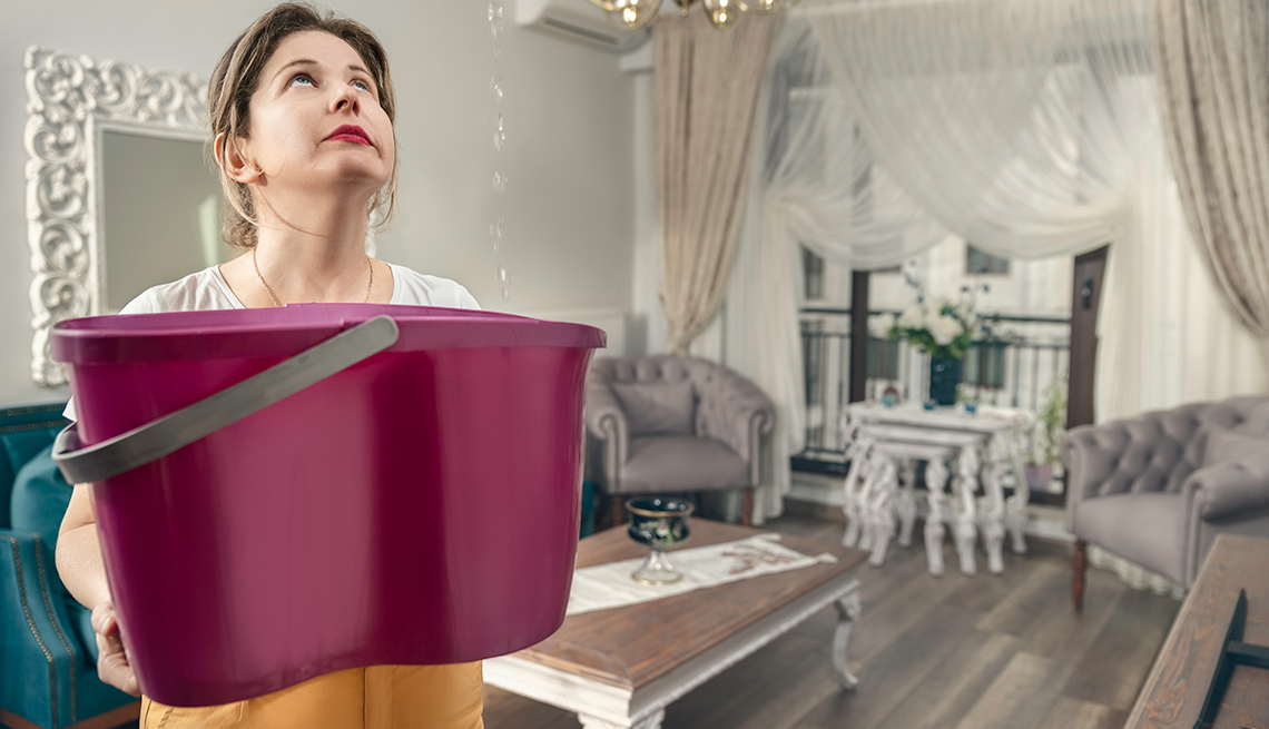 A woman looks up at a leaking living room ceiling while holding a bucket  to catch the water.