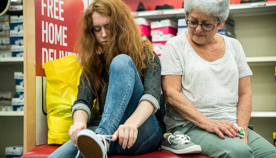 A grandmother and teenage granddaughter sit together on a bench in a shoe store as the teen trries on a pair of shoes.