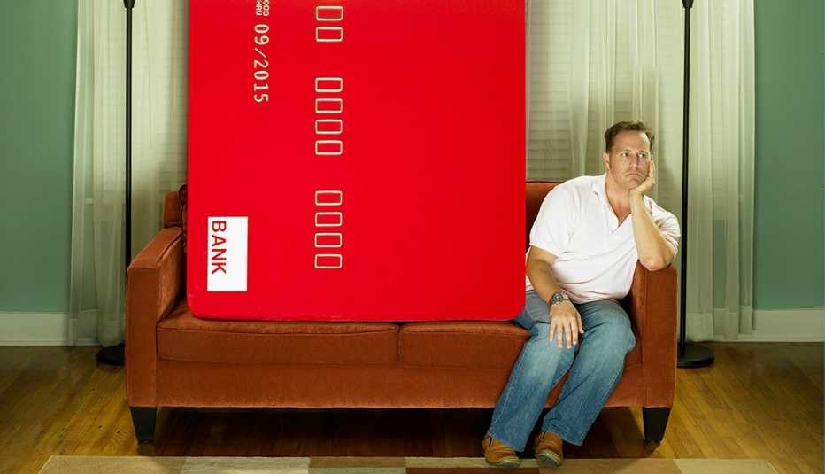 A man sits next to a huge credit card on a sofa. 
