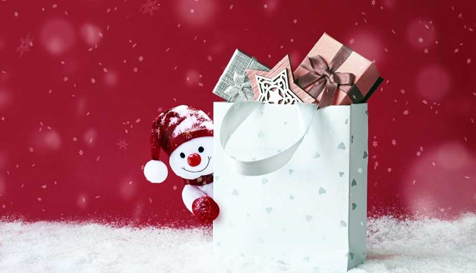 A Christmas snowman is shown peeking out from behind a bag of gifts which is sitting on snow against a festive red background.