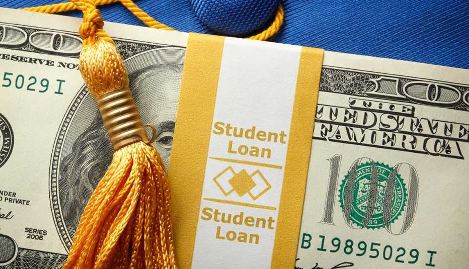 A stack of one hundred dollar bills in a money wrapper labeled "Student Loan" on top of a blue graduation cap.  A gold graduation tassel is draped over the stack of money.