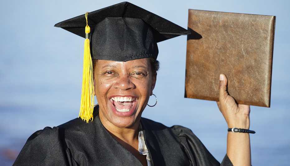 Happy portrait of a woman dressed in a graduation cap and gown holding up a diploma and smiling against a blue sky.