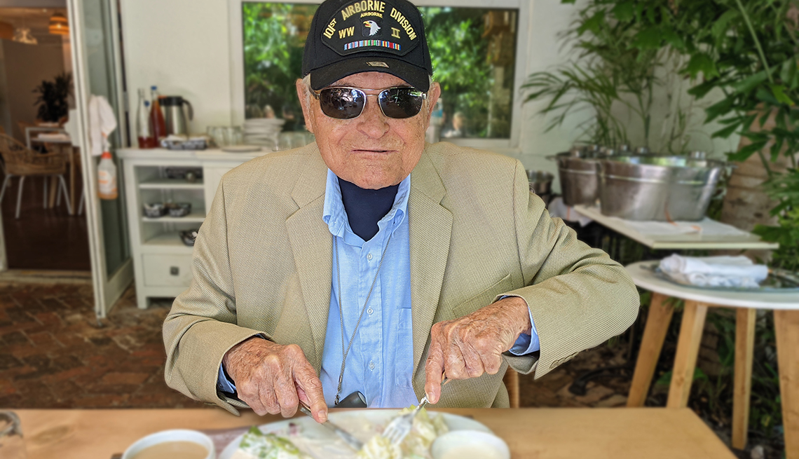 portrait of dapper 98-year-old investment guru Taylor Larimore seated to eat a meal; he is wearing sunglasses and a WWII US Army Airborne Division commemorative cap 