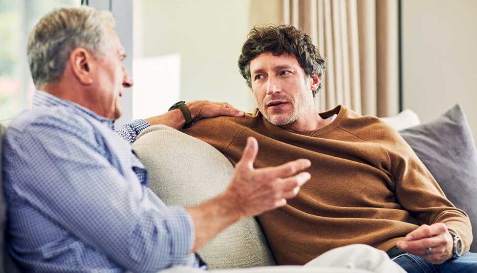 mature man and his senior father with hand on son's shoulder, sitting on the sofa at home and chatting
