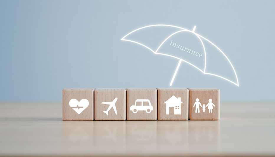 several wooden blocks on a table showing a health icon a plane a car a house and a family placed underneath a white outline of an umbrella that reads insurance