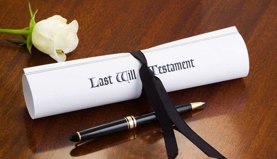 A will document tied with a black ribbon sits on a wood desk next to a white rose and a pen. 