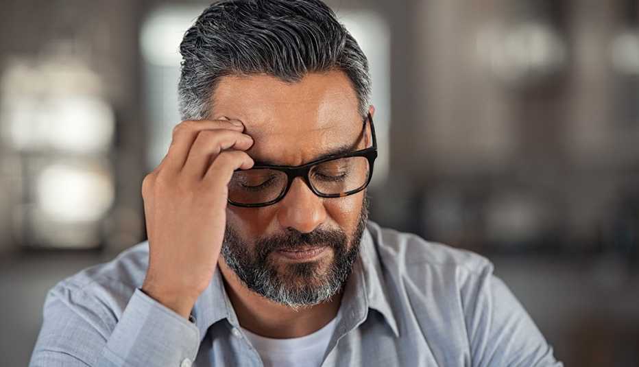 Man with hand on head sitting on couch at home. Close up face of stressed indian businessman wearing eyeglasses with eyes closed. 