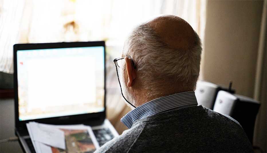 Over the shoulder view of a man who is half bald, wearing eyeglasses and using his laptop in the living room near window