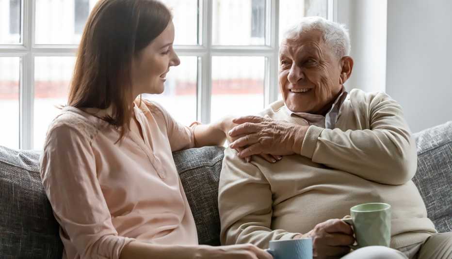 Young woman sitting on sofa with happy older retired 70s father, enjoying pleasant conversation with cup of coffee tea together in living room, mature parents and grown children communication.