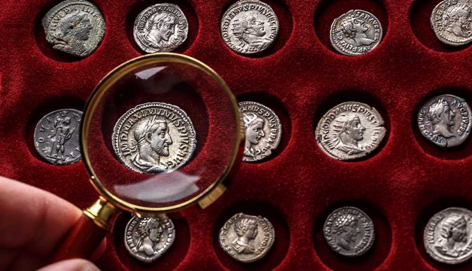 A collector holds a magnifying glass over an old denarius coin of the Roman Empire.