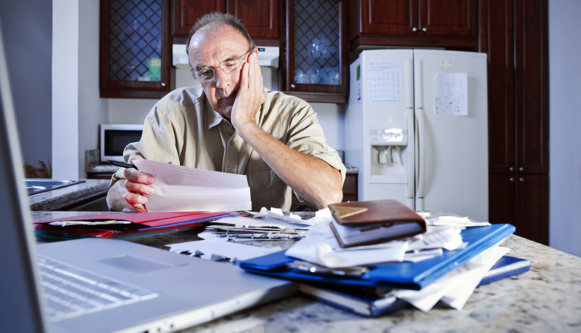 man sits at kitchen table with a pile of paperwork and laptop computer, looking stressed