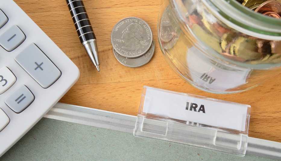 Desktop display of a paper file folder tab labeled "IRA" along with a money jar, quarters, a pen and a calculator