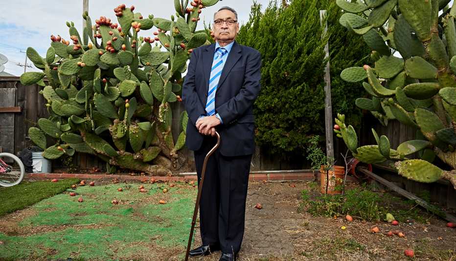 jose leaning on a cane standing in a yard with tall cactus plants and shrubbery behind him 