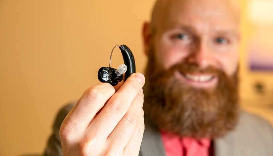 man blurred in background holding up an over-the-counter hearing aid in foreground