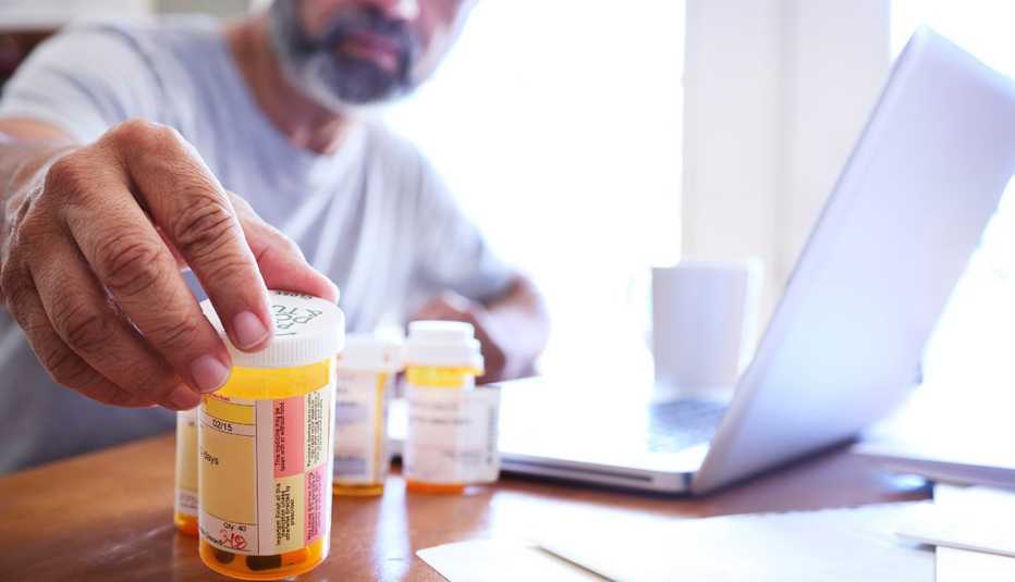 A Hispanic man in his late fifties reaches for one of his prescription medication bottles as he sits at his dining room table.  His laptop computer is open in front of him while sunlight filters in through the window behind him bathing the room with a sof