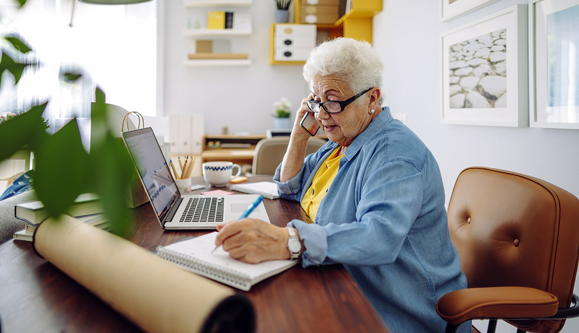 older female worker at home desk with mobile phone, laptop and notepad