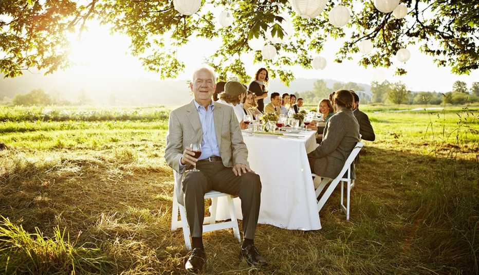 Senior man holding glass of wine sitting at head of banquet table outside in field friends and family seated behind