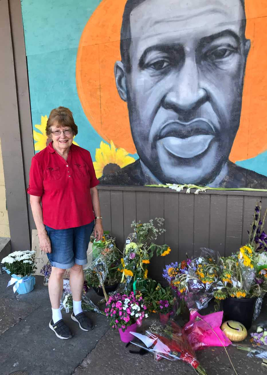 lois knowlton stands next to a memorial mural painted of george floyd that has piles of flowers laid underneath it