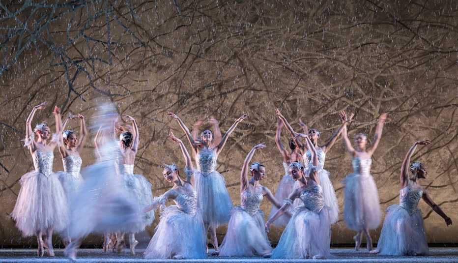 a dozen ballerinas in white and silver costumes dance on stage in front of a background of tree branches