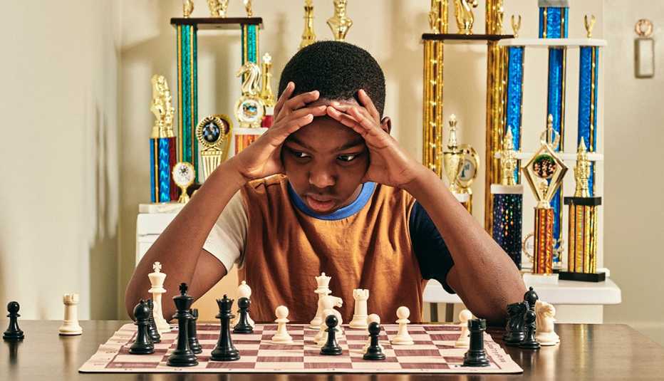 a young boy concentrates on a chessboard with trophies behind him