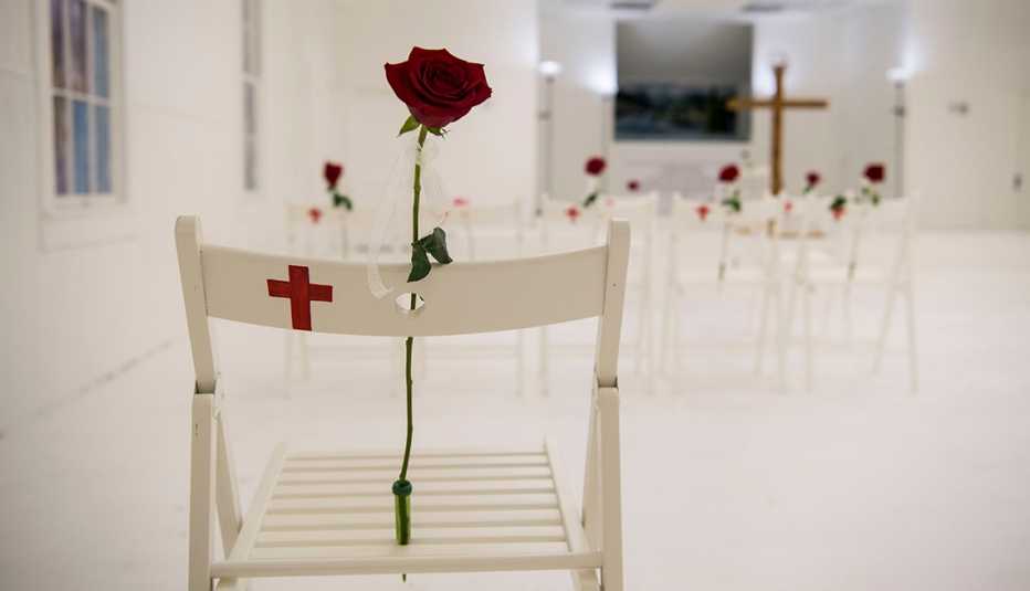 a memorial inside a church consists of twenty six empty white chars each with a red cross painted on it and a rose attached to its back