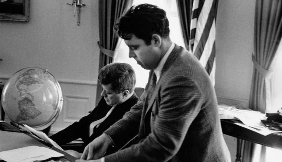 photographer jacques lowe standing next to president john f kennedy who is sitting at the desk in the oval office they are reviewing photographs together