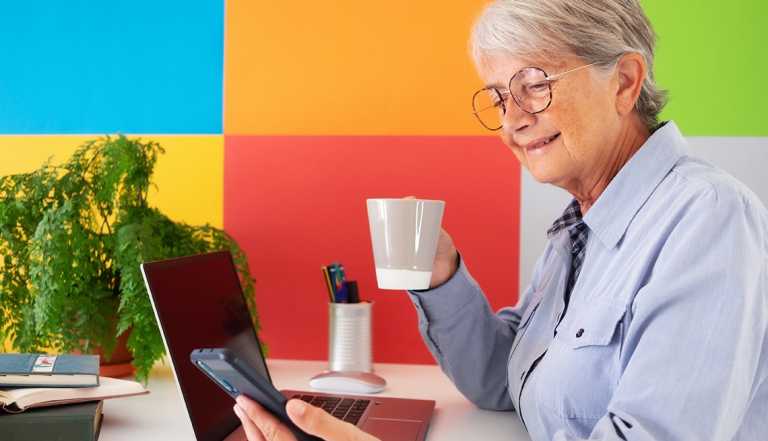A mature woman who is casually dressed and drinking coffee works remotely with her laptop and smartphone.