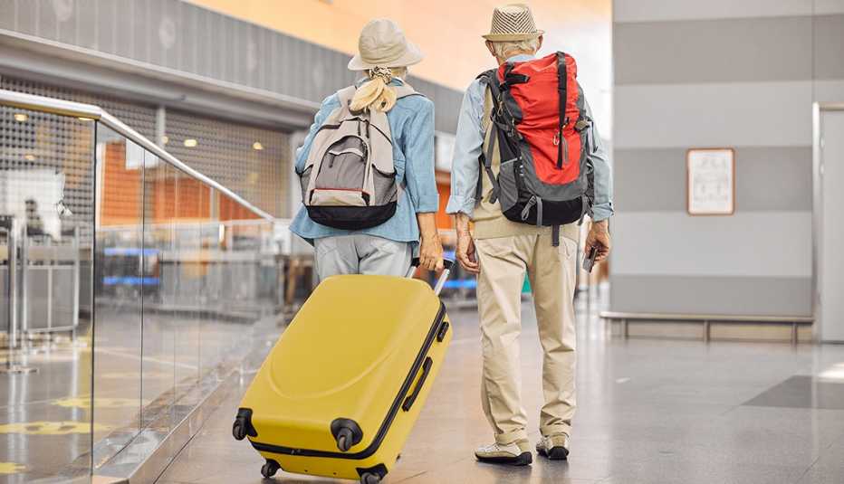 Back view of a retiree couple getting ready to depart at an airport with backpacks and a suitcase