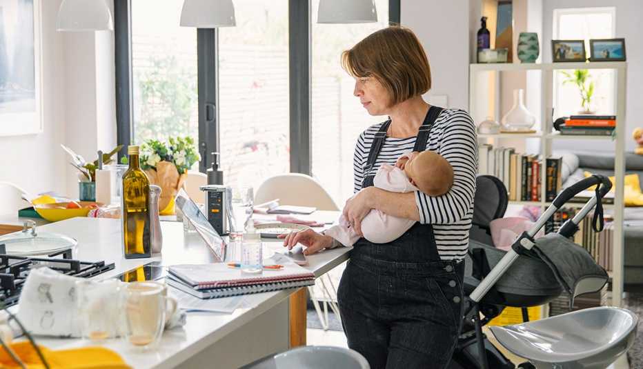 Older mother holding baby at home while working on laptop in kitchen