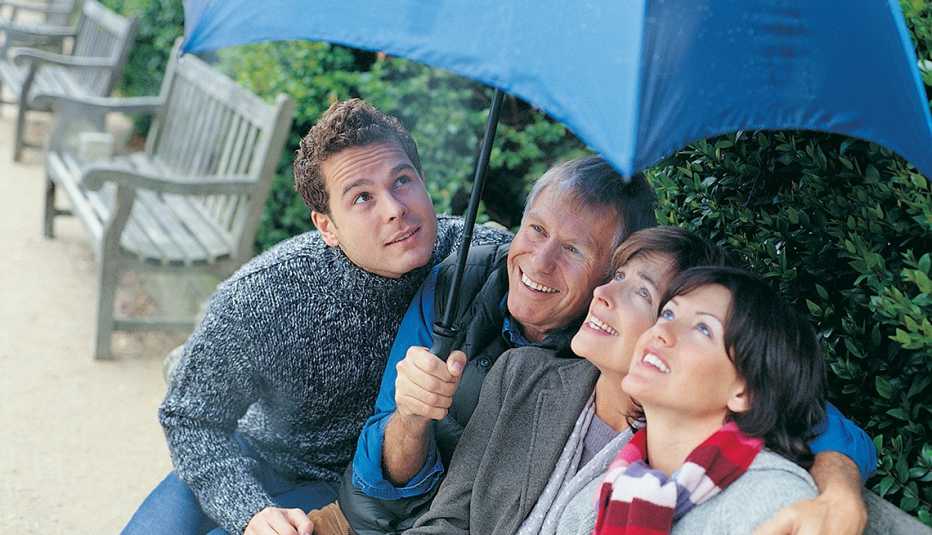 older couple and their adult children sitting on a park bench under an umbrella