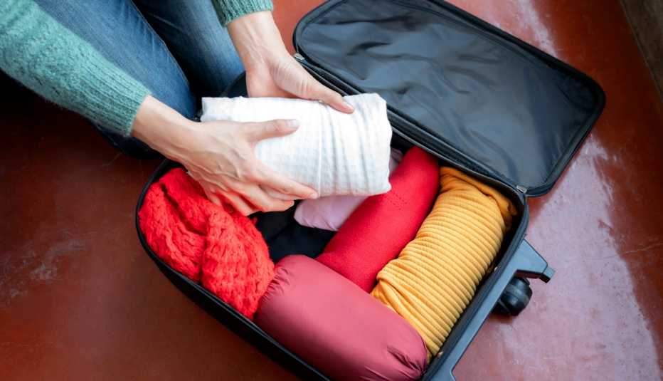 a woman packing rolled up sweaters and shirts into a carryon suitcase