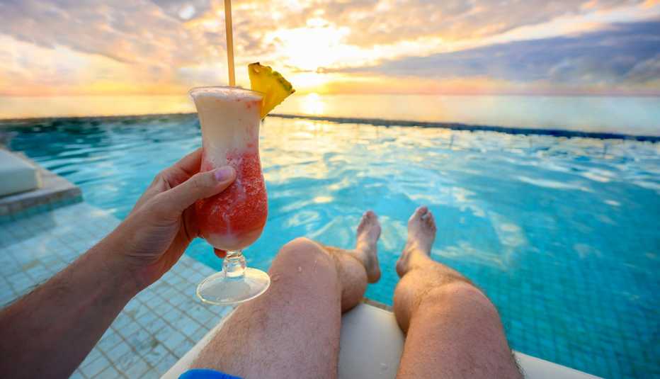View of a man sitting with his legs over an infinity pool on the ocean in Cancun. He's holing a colorful tropical frozen drink with a pineapple garnish