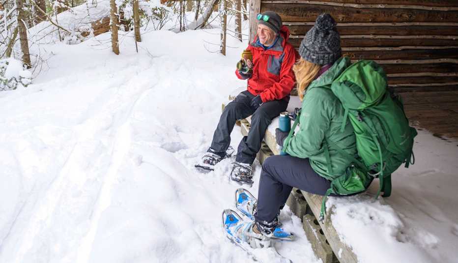 a man and a woman wearing coats and snowshoes take a break at a cabin