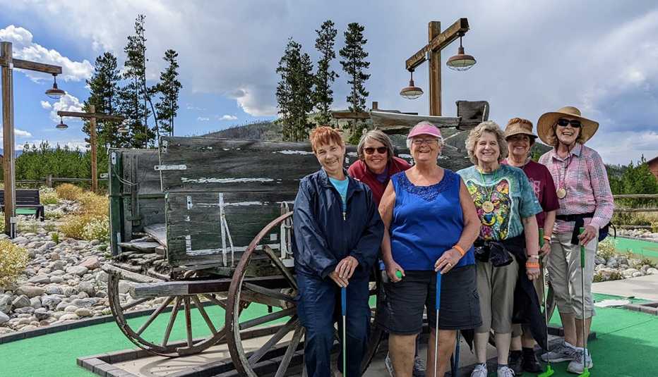 six senior women pose in front of an antique wagon on a miniature golf course in the mountains