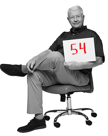 brian reid sits in an office desk chair holding a placard with the number 54 written on it