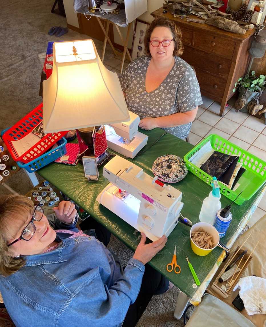 becky smith and her sister debbie cobb smile while looking up from their sewing machines