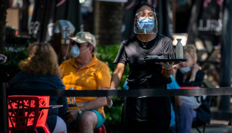 A waitress walks with a tray to a table while wearing a mask and face shield