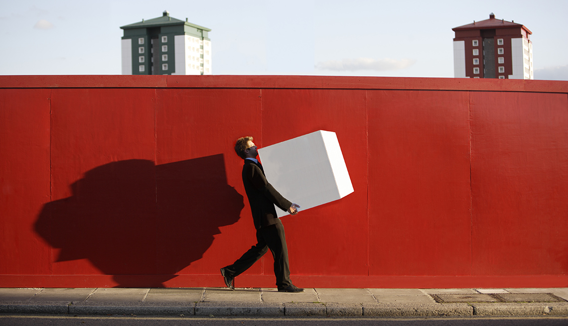 Man In Suit Holding Large White Box Walking Down Sidewalk With Red Wall Behind Him, AARP Work, Career Change, How To Negotiate A Killer Severance Package